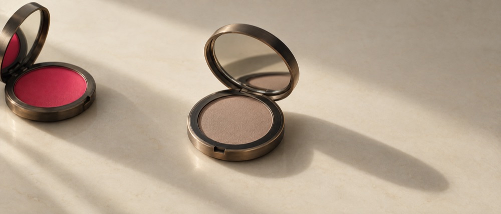 Photograph: two open powder compacts on porcelain, the loud red-pink one turned away, the muted taupe one centred in the light