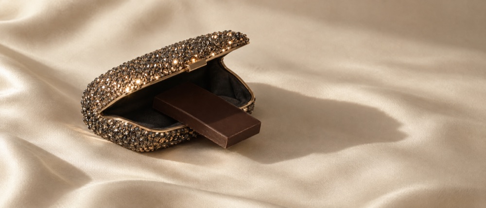 Photograph: a crystal-covered evening clutch open on ivory silk with a plain bar of dark chocolate resting inside it